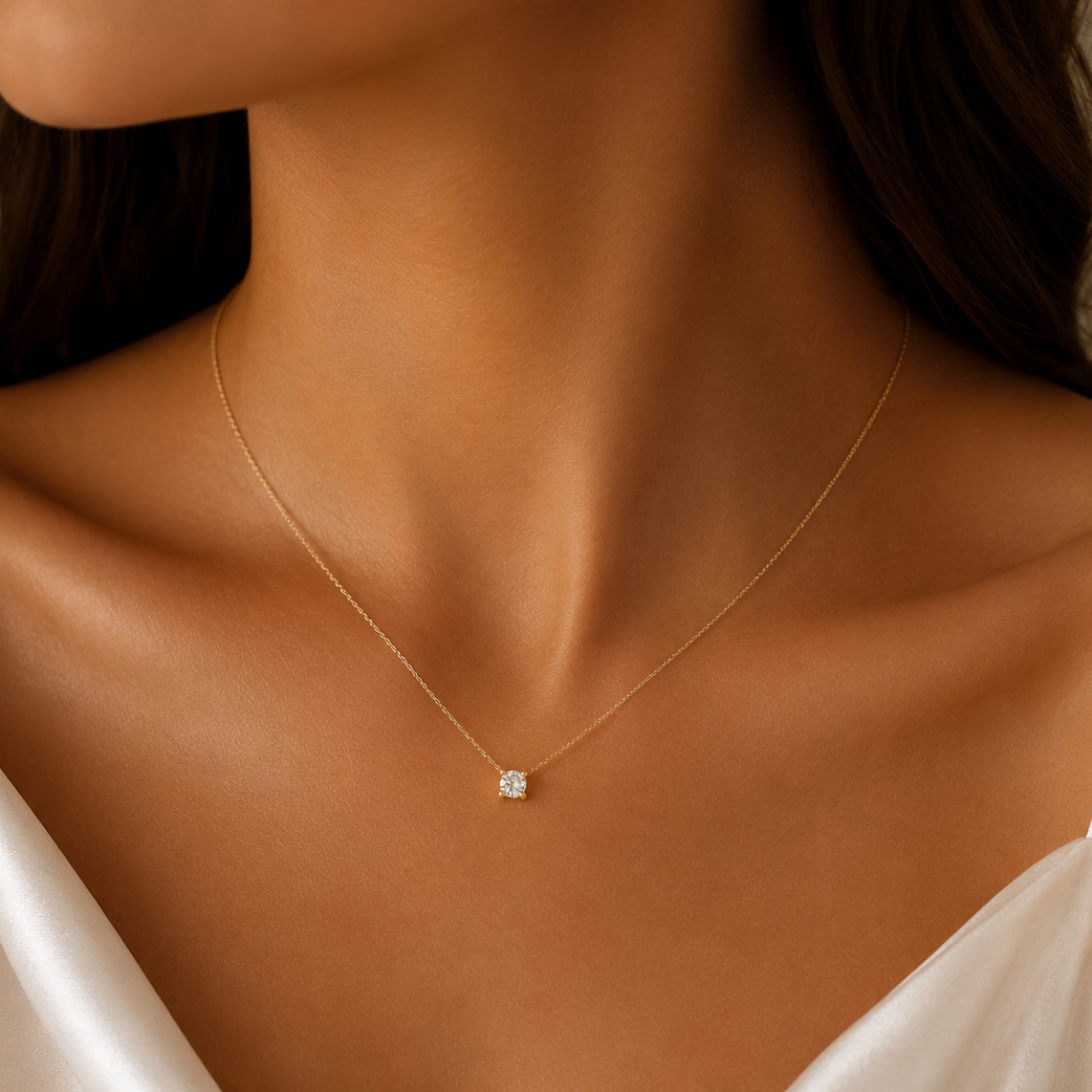 A close-up of a model wearing a delicate gold chain necklace with a small square-cut clear gemstone pendant, styled with a white satin camisole.