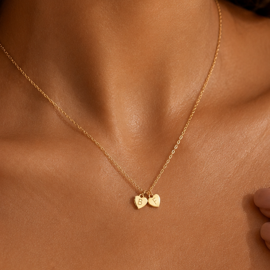 A delicate gold chain necklace worn on a model's neck, featuring two small interlocking heart charms, one engraved with the letter 'B'.