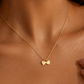 A delicate gold chain necklace worn on a model's neck, featuring two small interlocking heart charms, one engraved with the letter 'B'.