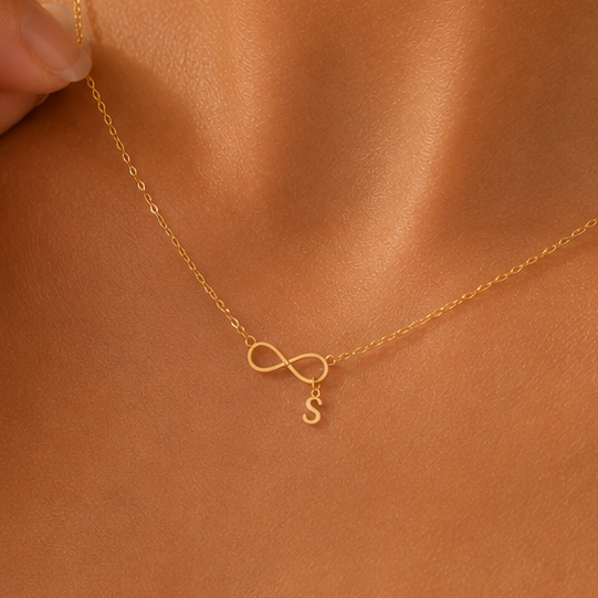 A close-up of a model wearing a delicate gold necklace with an infinity symbol and a small letter 'S' pendant, styled with a white satin camisole.