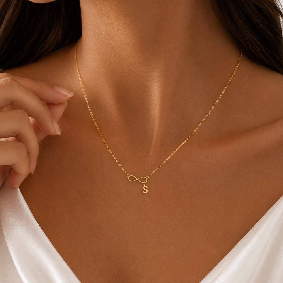 A close-up of a woman wearing a delicate gold necklace with an infinity symbol and a small initial 'S' pendant, styled with a white satin top.