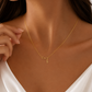 A close-up of a woman wearing a delicate gold necklace with an infinity symbol and a small initial 'S' pendant, styled with a white satin top.