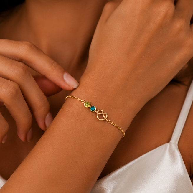A close-up of a delicate chain bracelet worn on a woman's wrist, with three additional color variants shown below: yellow gold, silver, and rose gold. Each bracelet features a small heart charm, a green oval stone, and a tiny blue oval stone.