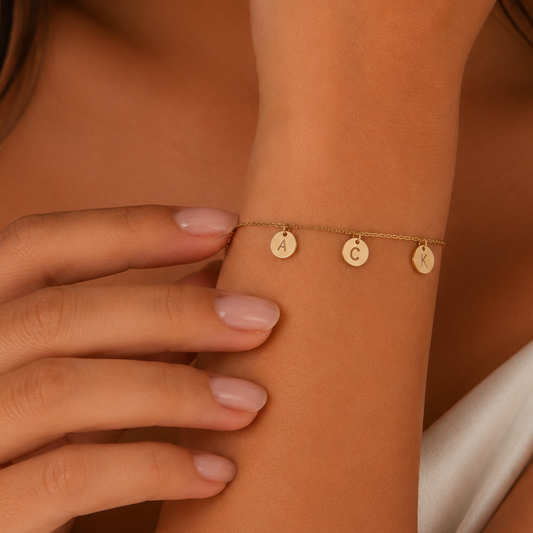 A delicate gold chain bracelet with three small circular disc charms, worn on a woman's wrist. The shot is a close-up lifestyle shot with warm, soft lighting and a white satin top in the background.