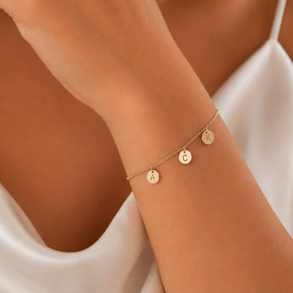 A delicate gold chain bracelet with three small round engraved disc charms worn on a woman's wrist. The shot is a close-up lifestyle crop showing the model's hand, shoulder, and white satin top.