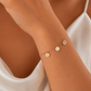 A delicate gold chain bracelet with three small round engraved disc charms worn on a woman's wrist. The shot is a close-up lifestyle crop showing the model's hand, shoulder, and white satin top.