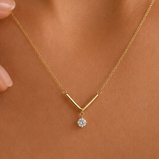 A delicate gold necklace with a V-shaped bar and a single round clear gemstone pendant, shown worn on a model's neck.