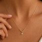 A close-up of a model's neck and chest wearing a delicate gold necklace with a V-shaped pendant and a small clear gemstone, with a hand gently touching the chain.