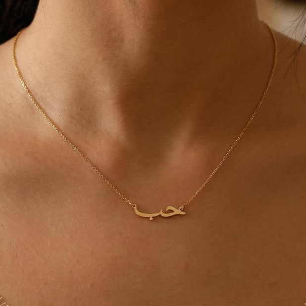 A delicate gold chain necklace with a small Arabic nameplate حب pendant in gold, worn by a model in a close-up shot.