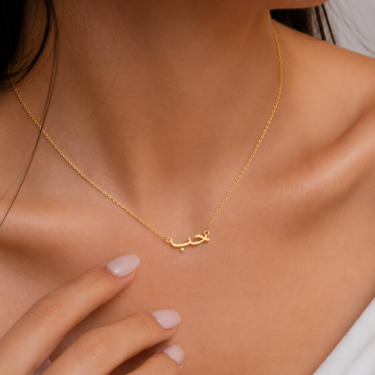 A delicate gold chain necklace with a small Arabic calligraphy pendant, shown worn on a model's neck with her hand resting near the chest.