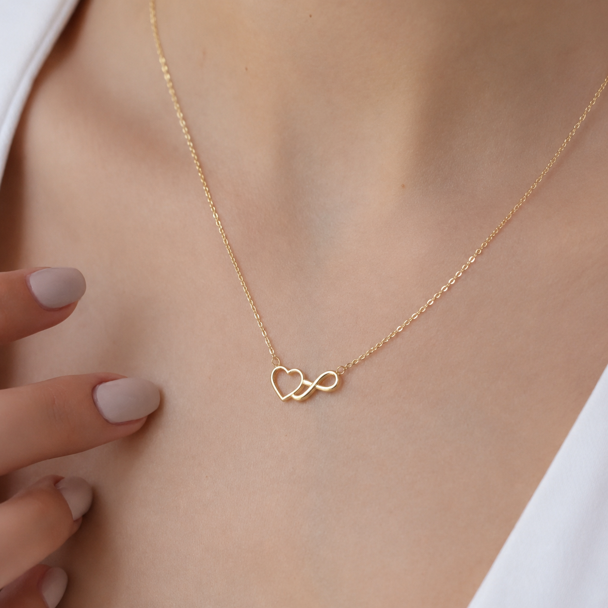 A delicate gold chain necklace with a double interlocking heart infinity pendant, shown worn on a model's neck with manicured nails visible on the side.